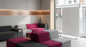 Collaborative Office Furniture - Professional 16:9 hero banner. Ultra high resolution. An architectural detail of a modern, inviting office space, focusing on a versatile modular seating arrangement in shades of grey and deep magenta (#E2087E). The area is bathed in a soft, strategic accent glow of #737373, highlighting the adaptable design and integrated technology ports subtly. A mobile whiteboard with faint, overlaid conceptual diagrams stands nearby, indicating dynamic teamwork. The setting reflects a prestigious, yet practical UK office environment, embodying Cost Cutters UK's expertise in creating functional, collaborative zones. Style: Editorial photography with a focus on clean lines and sophisticated design. Avoid: Groups around holographic tables, generic tech aesthetics, text overlays, cold blue tones, stock photo clichés.