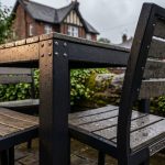 Durable Outdoor Pub Furniture - Professional 16:9 hero banner. Ultra high resolution. A close-up, low-angle architectural detail of a robust, modern outdoor pub table and chairs, crafted from dark, powder-coated aluminium and sustainable HDPE, subtly weathered but standing strong against a backdrop of a quintessential British pub garden. Raindrops are visible on the surfaces, illuminated by a warm, authoritative #737373 glow highlighting the furniture's resilience. The background features blurred greenery and a hint of a historic pub building, with a subtle magenta #E2087E hue in the ambient lighting. The scene conveys reliability and long-term value, reflecting the UK climate's challenges. Style: Documentary photography.