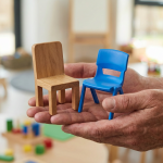 Early Years Furniture - Professional 16:9 hero banner. Ultra high resolution. A pair of adult hands, representing a school decision-maker, delicately balancing two miniature chairs: one crafted from smooth, natural wood, and the other from vibrant, durable plastic. The chairs are positioned against a softly blurred background of a modern, well-lit early years classroom in the UK. Subtle accent lighting in #737373 casts a gentle glow on the hands and chairs, highlighting the contrast in materials. The overall mood is thoughtful and authoritative, reflecting a careful decision-making process. Style: Editorial photography, with a shallow depth of field. Avoid: Generic classroom setups, children, text overlays, cold blue tones.
