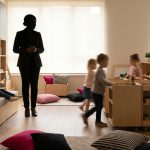 Multi-Functional Nursery Furniture - Professional 16:9 hero banner. Ultra high resolution. An authoritative, silhouetted figure, possibly a Headteacher or Nursery Manager, stands in a dynamically zoned, brightly lit nursery classroom. The figure is observing children (blurred in the background for focus) engaging with versatile, multi-functional furniture – perhaps a storage bench that doubles as a reading nook, or mobile units creating flexible activity zones. The room is designed with a sense of spaciousness and order, despite being compact. Accent lighting in #737373 highlights the furniture's adaptability, with elements of #E2087E and #000000 in the room's decor, suggesting a thoughtfully designed, engaging learning environment. Style: Editorial photography, conveying expertise and practical solutions. Avoid: Groups around holographic tables, generic tech aesthetics, text overlays, cold blue tones, stock photo clichés.