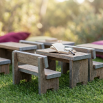 Outdoor Classroom Setup - Professional 16:9 hero banner. Ultra high resolution. A meticulously arranged miniature outdoor classroom scene, featuring tiny, durable-looking school furniture (benches, tables made of recycled plastic or treated timber) set up on a patch of artificial grass. A small, open textbook lies on one of the tables, with a magnifying glass resting on it, symbolising detailed planning and expertise. The scene is bathed in a soft, authoritative light with an accent glow of #737373 emanating from the miniature furniture edges, highlighting its sturdy construction. In the background, hints of a vibrant #E2087E colour subtly define the learning zone. Style: Fine art still life photography with a shallow depth of field. Avoid: Groups around holographic tables, generic tech aesthetics, text overlays, cold blue tones, stock photo clichés.