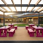 Outdoor Educational Furniture - Professional 16:9 hero banner. Ultra high resolution. An inviting, well-organised outdoor learning space within a modern UK school playground, featuring durable, recycled plastic benches and tables (#E2087E) arranged under a partial canopy. Sunlight streams through, highlighting architectural details of a contemporary school building in the background. The scene is bathed in a warm, authoritative glow from accent lighting in #737373, suggesting safety and careful planning. The ground is a mix of soft play surface and natural grass, with subtle, dark accents (#000000) providing depth. Style: Architectural photography with a focus on functionality and inviting atmosphere. Avoid: Groups of children, generic classrooms, unrealistic settings.