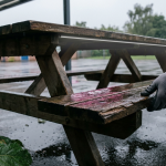 Outdoor Educational Furniture - Professional 16:9 hero banner. Ultra high resolution. A weathered yet well-maintained wooden picnic bench in a UK school playground, bathed in the soft, authoritative glow of #737373 accent lighting, hinting at care and longevity. Raindrops glisten on the surface, reflecting the #E2087E and #000000 tones in the puddles below, symbolising the challenging UK climate. A hand, gloved for protection, gently inspects a corner of the bench for splinters, emphasising safety and meticulous attention to detail. The scene conveys resilience and the quiet dedication of long-term asset care. Style: Editorial photography, with a focus on texture and natural light.