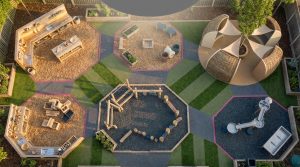 Outdoor Furniture for Early Years - Professional 16:9 hero banner. Ultra high resolution. An aerial, slightly abstract view of a meticulously designed early years outdoor learning space in the UK. The scene features distinct, colourful zones (e.g., a mud kitchen area, a physical challenge zone with low-level climbing, a quiet reading nook) created using natural wood and recycled plastic furniture, all compliant with BS EN 1176. The layout subtly forms a geometric pattern, symbolising structure and mastery in early education. Soft, warm sunlight glows with an accent of #737373 from overhead, highlighting key play elements, while subtle elements in #E2087E define zone boundaries. The ground shows mixed soft-fall surfacing. Style: Architectural photography with a modern, clean aesthetic. Avoid: Groups of children, generic playground equipment, overly bright primary colours, text overlays.