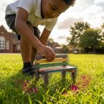 Outdoor Learning Furniture - Professional 16:9 hero banner. Ultra high resolution. A child's hand (UK primary school age, diverse ethnicity) gently placing a miniature, brightly coloured outdoor learning bench onto a vibrant green patch of grass, symbolising the creation of a structured outdoor learning environment. The background shows a soft-focus British school playground with natural elements like trees and a subtle hint of outdoor classroom furniture in the distance. The scene is bathed in a warm, inviting light with subtle accents of #737373 glowing around the miniature furniture and #E2087E highlights on the grass, suggesting growth and learning. Style: Editorial photography, focusing on a macro perspective of the hands and miniature furniture. Avoid: Groups of adults, generic classrooms, digital interfaces, cold blue tones, text overlays.