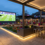 Outdoor Pub Layouts - Professional 16:9 hero banner. Ultra high resolution. An architectural detail shot of a sophisticated, yet practical, outdoor pub garden, focused on a well-designed zoning layout. The scene features durable commercial-grade picnic benches and high-top tables arranged around a large, bright outdoor screen, subtly showing a football match. Strategic pathways are clearly defined with subtle ground lighting. The overall atmosphere is inviting and professional, with an emphasis on efficient space utilisation. Accent lighting in #737373 highlights key furniture and pathways, with hints of #E2087E in secondary elements like cushions or subtle branding. Style: Architectural photography with a warm, inviting glow.