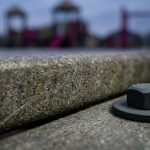 Outdoor School Furniture - Professional 16:9 hero banner. Ultra high resolution. A macro shot focusing on the intricate, weather-worn yet resilient surface of a recycled plastic (HDPE) plank, contrasted with a subtly textured powder-coated steel component, both illuminated by a precise (#737373) accent glow highlighting their durability. A faint, almost artistic hint of (#E2087E) suggests the vibrant life of a school playground in the background, out of focus. The composition emphasizes strength and longevity, with a subtle nod to sustainability. Style: Editorial still life photography, rich textures, shallow depth of field. Avoid: Generic school children, pristine new furniture, overtly digital interfaces.