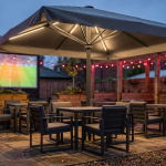 Weatherproof Pub Garden - Professional 16:9 hero banner. Ultra high resolution. A robust, stylish commercial outdoor umbrella, partially opened, with a subtle glow of #737373 light emanating from its interior ribs, casting dramatic shadows. Below it, high-quality, contract-grade pub garden furniture (dark grey powder-coated aluminium chairs and tables) stands ready. In the background, hints of a large outdoor screen showing a football match are visible, with warm, inviting string lights (#E2087E) draped overhead. The scene is set in a modern, well-maintained UK pub garden, showcasing readiness for any weather. Style: Editorial photography, focusing on texture and material quality. Avoid: Groups around holographic tables, generic tech aesthetics, text overlays, cold blue tones, stock photo clichés.