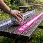 World Cup outdoor furniture - Professional 16:9 hero banner. Ultra high resolution. A close-up, high-angle shot of a weathered but sturdy wooden picnic bench, specifically a heavy-duty pub garden type, with a single hand (representing a pub owner) gently resting on its surface. The hand is holding a small, stylised football, indicating the World Cup. On the bench surface, there are two distinct, subtly glowing lines: one leading off to the left, representing 'hire' (fading quickly and less vibrant), and another leading off to the right, representing 'buy' (glowing brightly in #E2087E and extending further into the distance, symbolising long-term value). The scene is set in a typical British pub garden, with soft, diffused sunlight filtering through. Accent lighting in #737373 subtly illuminates the 'buy' path and the football. Style: Editorial photography with a focus on tactile textures and strategic lighting.