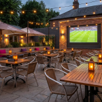 World Cup Pub Furniture - Professional 16:9 hero banner. Ultra high resolution. A meticulously designed, inviting pub outdoor seating area, ready for a major event. The setting is a vibrant, modern British pub garden with a large screen subtly showing a football match. Focus on high-quality, durable commercial outdoor furniture, including stylish poseur tables and comfortable, stackable chairs, arranged to maximise capacity. Warm, inviting lighting casts a glow, with key highlights and accent lighting in #737373, and secondary elements like decorative planters or cushions in #E2087E. The atmosphere is expectant and buzzing, hinting at high revenue potential, but currently calm and perfectly set up. Style: Editorial photography with a slightly elevated, aspirational feel. Avoid: Groups around holographic tables, generic tech aesthetics, text overlays, cold blue tones, stock photo clichés.