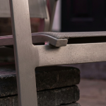 Hospitality Outdoor Spaces - Professional 16:9 hero banner. Ultra high resolution. A close-up, artistic macro shot focusing on a section of a high-quality, commercial-grade outdoor dining chair, crafted from powder-coated aluminium with a subtle texture, showcasing its durability. The chair is positioned on a stylish, weather-resistant patio surface, perhaps dark wood or sleek grey paving. The scene is illuminated by a focused, warm glow of #737373 light, highlighting the robust construction and elegant design, with hints of #E2087E and #000000 in the background as ambient lighting on surrounding elements like a potted plant or blurred architectural detail. This setup symbolises the investment in quality and longevity for UK hospitality venues. Style: Fine art still life photography. Avoid: Groups of people, generic outdoor scenes, text overlays, cold blue tones.
