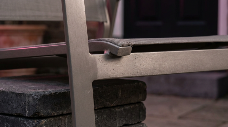 Hospitality Outdoor Spaces - Professional 16:9 hero banner. Ultra high resolution. A close-up, artistic macro shot focusing on a section of a high-quality, commercial-grade outdoor dining chair, crafted from powder-coated aluminium with a subtle texture, showcasing its durability. The chair is positioned on a stylish, weather-resistant patio surface, perhaps dark wood or sleek grey paving. The scene is illuminated by a focused, warm glow of #737373 light, highlighting the robust construction and elegant design, with hints of #E2087E and #000000 in the background as ambient lighting on surrounding elements like a potted plant or blurred architectural detail. This setup symbolises the investment in quality and longevity for UK hospitality venues. Style: Fine art still life photography. Avoid: Groups of people, generic outdoor scenes, text overlays, cold blue tones.