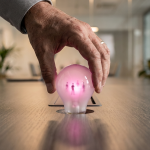 employee feedback - Professional 16:9 hero banner. Ultra high resolution. A close-up, editorial photography shot of a single, experienced hand (representing a manager or facilities professional) holding a stylised, glowing lightbulb. The lightbulb is translucent with a subtle internal glow in #E2087E, and contains tiny, abstract human figures (representing employees) inside, suggesting 'ideas' or 'feedback'. The hand is carefully placing this 'feedback lightbulb' into a slot on a sleek, dark office desk surface, which subtly reflects #737373 accent lighting. The background is a softly blurred, modern office interior, hinting at ergonomic furniture and collaborative spaces. The overall mood is authoritative and insightful, emphasising the value of employee input in shaping the physical workspace. Style: Editorial photography. Avoid: Groups around holographic tables, generic tech aesthetics, text overlays, cold blue tones, stock photo clichés.