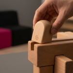 EYFS Compliance - Professional 16:9 hero banner. Ultra high resolution. A macro shot focusing on a meticulously crafted, child-safe wooden building block being placed perfectly into a larger, intricate wooden structure. The block, smooth and rounded, symbolises a key component of a compliant early years environment. Subtle accent lighting in #737373 casts a protective glow over the scene, highlighting the precision and care. In the background, blurred elements of purpose-built, certified educational furniture in #E2087E and #000000 tones provide context. The overall mood is one of meticulous attention to detail and foundational safety. Style: Editorial still life photography, with a shallow depth of field.