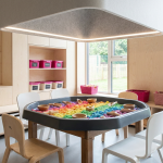 Messy Play - Professional 16:9 hero banner. Ultra high resolution. An architectural detail of a modern, well-organised early years classroom or nursery, focusing on a dedicated 'messy play zone'. The area features a robust, clean Tuff Tray filled with colourful, safe sensory materials (e.g., kinetic sand, coloured rice), surrounded by child-sized, easy-to-clean furniture. Bright, natural light illuminates the scene, with an accent glow of #737373 highlighting the play area, and hints of #E2087E in subtle educational elements. The scene conveys a sense of calm, structured learning and playful exploration, dispelling chaos. Style: Editorial photography, clean lines, inviting atmosphere. Avoid: Groups of children, overly chaotic scenes, generic classroom setups.