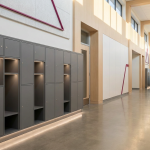 Mobile Phone Bans in Schools - Professional 16:9 hero banner. Ultra high resolution. A pristine, modern school corridor, designed with clean lines and natural light, leading towards a classroom door. The focus is on a sleek, multi-compartment locker unit, made of durable grey metal, subtly glowing with a #737373 accent light from within some compartments, hinting at secure storage. The background hints at the vibrant #E2087E through subtle architectural elements or distant wall art. The scene evokes order, security, and a focused learning environment, reflecting the authoritative and professional brand personality. Style: Architectural photography with a clean, editorial feel. Avoid: Groups of students, visible mobile phones, text overlays, generic tech aesthetics, cold blue tones.