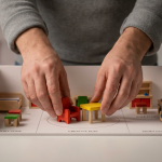 Nursery Room Layouts - Professional 16:9 hero banner. Ultra high resolution. A pair of adult hands, gently and meticulously arranging miniature, brightly coloured wooden furniture pieces within a scaled-down, pristine white model of a nursery room. The model clearly shows distinct 'zones' marked on the floor, representing different EYFS learning areas. Soft, focused accent lighting in #737373 highlights the hands and the intricate details of the miniature furniture, casting subtle shadows. The background is a clean, minimalist studio setting, with a hint of #E2087E in the soft-focused distance. The overall atmosphere is one of careful planning, precision, and a nurturing approach to early years education, reflecting Cost Cutters UK's expertise in design and installation. Style: Detailed macro photography with a shallow depth of field. Avoid: Groups around holographic tables, generic tech aesthetics, text overlays, cold blue tones, stock photo clichés.