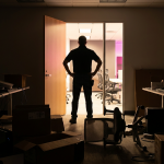 Office Design Mistakes - Professional 16:9 hero banner. Ultra high resolution. A solitary, experienced facilities manager, silhouetted slightly, stands in a dimly lit, chaotic office space that visually represents common 'office design mistakes'. The foreground shows a tangled mess of wires, an uncomfortable, generic office chair overturned, and a stack of misaligned boxes, all cast in dramatic shadows. Behind them, through a doorway, a glimpse of a perfectly organised, brightly lit, modern office with ergonomic furniture and clear pathways is visible, illuminated by a warm #737373 accent glow and hints of #E2087E. The manager looks towards this ideal space, hands on hips, conveying a sense of authority and problem-solving. Style: Documentary photography with a focus on contrasting light and shadow. Avoid: Groups around holographic tables, generic tech aesthetics, text overlays, cold blue tones, stock photo clichés.