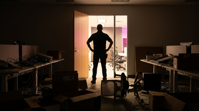 Office Design Mistakes - Professional 16:9 hero banner. Ultra high resolution. A solitary, experienced facilities manager, silhouetted slightly, stands in a dimly lit, chaotic office space that visually represents common 'office design mistakes'. The foreground shows a tangled mess of wires, an uncomfortable, generic office chair overturned, and a stack of misaligned boxes, all cast in dramatic shadows. Behind them, through a doorway, a glimpse of a perfectly organised, brightly lit, modern office with ergonomic furniture and clear pathways is visible, illuminated by a warm #737373 accent glow and hints of #E2087E. The manager looks towards this ideal space, hands on hips, conveying a sense of authority and problem-solving. Style: Documentary photography with a focus on contrasting light and shadow. Avoid: Groups around holographic tables, generic tech aesthetics, text overlays, cold blue tones, stock photo clichés.