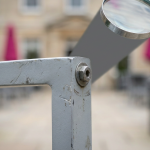Outdoor Hospitality Furniture - Professional 16:9 hero banner. Ultra high resolution. A close-up, macro shot of a weathered, yet structurally sound, corner joint of a high-quality outdoor hospitality chair. The joint showcases resilient, powder-coated aluminium with subtle signs of long-term use, but no rust or degradation. A magnifying glass hovers over the joint, with a focused beam of light in brand accent colour #737373 highlighting the detail, symbolising thorough inspection and debunking myths. The background is a subtly blurred outdoor setting, perhaps a sophisticated hotel patio or school courtyard in the UK, with hints of magenta #E2087E and black #000000 in the distance. Style: Detailed editorial photography, emphasising material integrity. Avoid: Groups around tables, generic tech, text overlays.