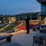 Outdoor Hospitality Planning - Professional 16:9 hero banner. Ultra high resolution. An authoritative, silhouetted figure, perhaps a facilities manager or procurement officer, stands confidently on a modern outdoor patio, overlooking a thoughtfully designed commercial outdoor space. The setting is a contemporary UK business or education campus, featuring durable, stylish outdoor furniture made from recycled plastic and powder-coated aluminium. The scene is illuminated by striking accent lighting in #737373, casting a warm glow on the furniture and highlighting architectural elements, with subtle hints of #E2087E in planters or decorative features. The overall mood is professional and forward-thinking, emphasising strategic planning and long-term value. Style: Architectural photography with a focus on design and functionality. Avoid: Groups around holographic tables, generic tech aesthetics, text overlays, cold blue tones, stock photo clichés.