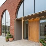 School Nursery Expansion - Professional 16:9 hero banner. Ultra high resolution. Architectural detail of a modern, inviting UK school nursery entrance, with large, child-friendly windows and a bright, welcoming door. The building material is a warm, red brick, indicative of a classic British school, but with contemporary, clean lines. Soft, natural light streams through, accented by a subtle #737373 glow around the entrance, hinting at new opportunities. A hint of vibrant #E2087E can be seen in a playful, abstract mural nearby. The scene conveys trust, growth, and a professional, authoritative tone. Style: Editorial architectural photography. Avoid: Groups around holographic tables, generic tech aesthetics, text overlays, cold blue tones, stock photo clichés.