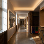 School Space Utilisation - Professional 16:9 hero banner. Ultra high resolution. A wide-angle, architectural detail shot inside a modern, yet established, UK school corridor. The perspective is looking down a long, well-lit corridor with subtle indications of flexible learning zones (e.g., a high counter with stools in a recess, mobile storage units). The space feels organised and purposeful. Sunlight streams through a large window, highlighting dust motes, with a subtle #737373 accent glow emanating from integrated lighting fixtures or digital displays on the walls. A hint of #E2087E could be seen in the colour of a plant pot or a subtle design element, with #000000 providing depth in shadows. Style: Editorial architectural photography. Avoid: Groups around holographic tables, generic tech aesthetics, text overlays, cold blue tones, stock photo clichés.