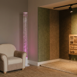 Sensory Play Environments - Professional 16:9 hero banner. Ultra high resolution. Architectural detail of a thoughtfully designed, inclusive early years classroom corner, bathed in a soft, inviting glow of #737373 light. Focus on a 'calm-down corner' with plush, child-friendly seating and a subtle bubble tube glowing with #E2087E light, suggesting both stimulation and serenity. Textured wall panels in muted tones, with a small, focused spotlight of #000000 illuminating a tactile board. The scene evokes a sense of safety, learning, and thoughtful design for young children. Style: Editorial photography with a warm, documentary feel. Avoid: Overly bright primary colours, cluttered spaces, groups of children, generic office settings.