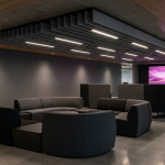 The Future of Work - Professional 16:9 hero banner. Ultra high resolution. A sophisticated, contemporary office interior, focusing on an architectural detail – perhaps a sleek, ergonomic breakout furniture zone with adaptable, modular seating in dark greys and blacks. The space is subtly illuminated by accent lighting in #737373, highlighting the clean lines and flexible nature of the furniture. A soft glow of #E2087E emanates from a distant digital display showing abstract data visualisations, symbolising innovation and foresight. The atmosphere is authoritative yet inviting, reflecting a human-centric design approach. Style: Editorial architectural photography. Avoid: Groups around holographic tables, generic tech aesthetics, text overlays, cold blue tones, stock photo clichés.
