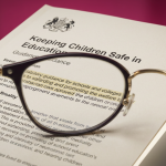 UK Education Legislation - Professional 16:9 hero banner. Ultra high resolution. A finely detailed, slightly elevated macro shot focusing on a section of an open, official-looking UK government guidance document, perhaps 'Keeping Children Safe in Education' or 'School Premises Regulations', with key phrases subtly highlighted. A pair of professional spectacles rests on the page, symbolising diligent review and expertise. The scene is illuminated by an accent glow of #737373 emanating from beneath the document, casting a professional, authoritative light. Subtle magenta (#E2087E) hints appear in the background, perhaps as a soft, out-of-focus wall colour. The overall mood is serious and focused, reflecting the importance of compliance for educational institutions. Style: Editorial still life photography. Avoid: Groups around holographic tables, generic tech aesthetics, text overlays, cold blue tones, stock photo clichés.