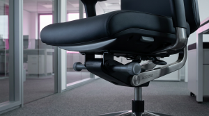 Advanced Ergonomics - Professional 16:9 hero banner. Ultra high resolution. A close-up, architectural detail shot of a sophisticated, high-end ergonomic office chair, focusing on its intricate adjustment mechanisms. The chair is positioned in a sleek, modern UK office interior with subtle light reflections. The shot highlights the precision engineering of the chair's components, with a strong accent glow of #737373 emanating from beneath the seat and along adjustment levers, contrasting with the chair's dark upholstery and the #E2087E subtle backlighting of a distant window. The overall mood is authoritative and technical, showcasing the chair as a piece of advanced engineering. Style: Editorial photography with a focus on product design.