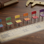 Classroom Chairs by Age - Professional 16:9 hero banner. Ultra high resolution. A close-up, macro shot of a meticulously arranged display of miniature, colour-coded classroom chairs (representing Size Marks 1-6), with each chair precisely aligned to an educational ruler or blueprint that clearly indicates age ranges and heights. The scene is set on a polished, dark wooden desk, suggesting a school procurement manager's organised workspace. A soft, authoritative glow in #737373 emanates from behind the chairs, highlighting their forms and the precision of the arrangement, while subtle accents of #E2087E appear in the chair colours. The overall mood is one of detailed expertise and careful planning. Style: Still life photography, editorial. Avoid: Groups around holographic tables, generic tech aesthetics, text overlays, cold blue tones, stock photo clichés.