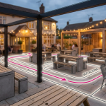 Designing Pub Gardens - Professional 16:9 hero banner. Ultra high resolution. An intricately designed blueprint overlay of a modern pub garden layout, featuring distinct zones for dining and socialising, with commercial-grade furniture outlines. The blueprint is subtly illuminated by a #737373 accent glow, highlighting key areas and strategic planning elements. A strong #E2087E line delineates profitable flow paths, with subtle #000000 accents for structural elements. The background shows a soft-focus, inviting pub garden setting with durable furniture. Style: Architectural photography with a conceptual overlay. Avoid: Groups around holographic tables, generic tech aesthetics, text overlays, cold blue tones, stock photo clichés.