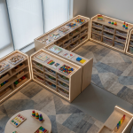 Early Years Storage Solutions - Professional 16:9 hero banner. Ultra high resolution. An aerial view of a meticulously organised early years classroom. Low-level wooden shelving units, each filled with colourful, accessible learning resources in clear trays, form a structured yet inviting environment. Soft, natural light streams in, with subtle accents of #737373 glowing around key storage areas, highlighting their accessibility and organisation. The floor features a playful, child-friendly mat with muted geometric patterns. A few small, age-appropriate educational toys are neatly arranged, demonstrating a calm and purposeful learning space. The overall mood is authoritative, clean, and highly functional, reflecting Cost Cutters UK's expertise in educational furniture. Style: Architectural photography with a focus on interior design and functionality. Avoid: Groups of people, cluttered spaces, or overtly playful elements that detract from the professional tone.