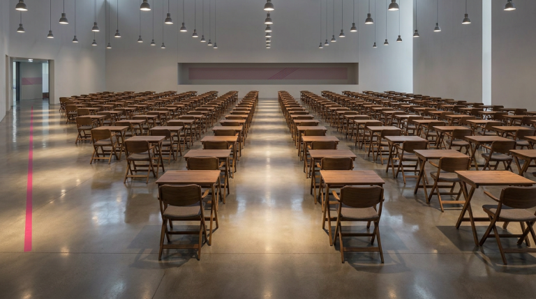 Exam Season Setup - Professional 16:9 hero banner. Ultra high resolution. A meticulously arranged, empty exam hall seen from a slightly elevated, wide-angle perspective. The scene focuses on rows of perfectly spaced, sturdy folding exam desks and ergonomic chairs, illuminated by a warm, focused glow of #737373 light emanating from ceiling fixtures, highlighting the organised precision. A subtle hint of #E2087E could be seen in distant wall art or a floor marker. The atmosphere is calm, prepared, and authoritative, ready for students. The floor gleams, reflecting the light, emphasising cleanliness and order. Style: Editorial architectural photography.