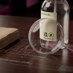 Janitorial Supplies UK - Professional 16:9 hero banner. Ultra high resolution. A meticulously arranged still life composition on a dark, polished surface. The central focus is a pristine, clear magnifying glass, its lens perfectly highlighting a small, detailed label on an 'eco-friendly' cleaning product bottle, showing UK compliance symbols. Surrounding it are miniature, stylised representations of key elements: a stack of neatly folded paper towels, a small, concentrated cleaning solution bottle, and a subtle blueprint overlay indicating a floor plan of a school or office. The scene is bathed in a dramatic accent glow of #737373, with subtle hints of #E2087E in the background, creating a sense of precision and strategic analysis. Style: Detailed still life photography with a shallow depth of field. Avoid: Groups around holographic tables, generic tech aesthetics, text overlays, cold blue tones, stock photo clichés.