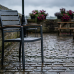 Outdoor Hospitality Seating - Professional 16:9 hero banner. Ultra high resolution. A solitary, weathered but robust outdoor cafe chair, made of powder-coated aluminium and treated hardwood, stands firm on a damp, historic UK cobblestone patio. Raindrops glisten on its surface, reflecting a subtle #737373 accent glow, highlighting its durability against the UK's unpredictable weather. In the background, hints of an inviting but empty pub garden, with a touch of #E2087E in flowering planters, evoke the potential for transformation. The scene conveys resilience and long-term value, focusing on the chair's ability to withstand harsh conditions. Style: Documentary photography, with a slightly dramatic, authoritative mood.