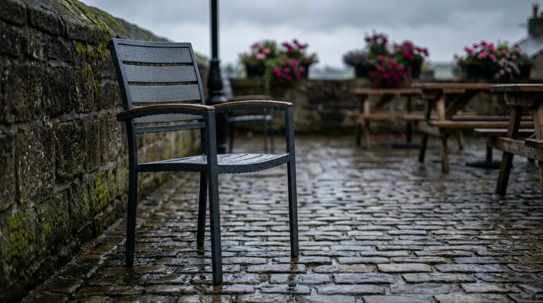 Outdoor Hospitality Seating - Professional 16:9 hero banner. Ultra high resolution. A solitary, weathered but robust outdoor cafe chair, made of powder-coated aluminium and treated hardwood, stands firm on a damp, historic UK cobblestone patio. Raindrops glisten on its surface, reflecting a subtle #737373 accent glow, highlighting its durability against the UK's unpredictable weather. In the background, hints of an inviting but empty pub garden, with a touch of #E2087E in flowering planters, evoke the potential for transformation. The scene conveys resilience and long-term value, focusing on the chair's ability to withstand harsh conditions. Style: Documentary photography, with a slightly dramatic, authoritative mood.