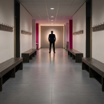 School Cloakroom Storage - Professional 16:9 hero banner. Ultra high resolution. An authoritative, silhouetted figure, possibly a School Business Manager, stands confidently at the end of a brightly lit, impeccably organised school cloakroom corridor. The corridor features modern, durable Solid Grade Laminate benches and wall-mounted coat hooks, with clear, wide walkways. The scene is illuminated by an accent glow of #737373, highlighting the clean lines and order, with secondary elements in #E2087E. The overall atmosphere is one of efficiency and safety, reflecting Cost Cutters UK's expertise in educational furniture solutions. Style: Editorial photography. Avoid: Groups around holographic tables, generic tech aesthetics, text overlays, cold blue tones, stock photo clichés.