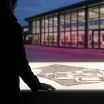 school facilities management - Professional 16:9 hero banner. Ultra high resolution. An authoritative, silhouetted figure, subtly representing a school business manager, stands in the foreground, facing a strategic blueprint of a school campus laid out on a large, illuminated table. The blueprint shows a mix of 'hard' elements like buildings and 'soft' elements like landscaped grounds, highlighted with a #737373 accent glow. The background features a subtly blurred, modern school building exterior at dusk, with some interior lights casting a #E2087E hue. The mood is strategic and forward-thinking. Style: Editorial photography, with a focus on depth and professional atmosphere. Avoid: Groups around holographic tables, generic tech aesthetics, text overlays, cold blue tones, stock photo clichés.