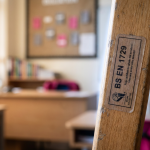 school furniture regulations UK - Professional 16:9 hero banner. Ultra high resolution. A finely detailed macro shot of a British Standard (BS EN) certification label, prominently displayed on a small, sturdy wooden school chair leg. The label is crisp and legible, highlighting the 'BS EN 1729' text. The background is a subtly blurred, warm-toned classroom environment with natural light filtering in, suggesting safety and order. A soft, authoritative glow of #737373 emanates from behind the label, with subtle accents of #E2087E and #000000 in the background elements, indicating precision and adherence to standards. Style: Documentary photography, focus on texture and detail.