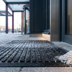Workplace Winter Safety - Professional 16:9 hero banner. Ultra high resolution. A close-up, architectural detail shot of a modern, commercial building entrance in the UK during a crisp winter morning. The focus is on a high-traction, heavy-duty outdoor mat covered with a light dusting of snow, with a pile of salt crystals subtly visible nearby. A subtle glow from accent colour #737373 highlights the mat's texture and the building's clean lines, suggesting preparedness and safety. The background shows a hint of a well-gritted path leading towards the entrance, with structural elements subtly outlined in primary colour #E2087E. The overall mood is authoritative and reassuring. Style: Architectural photography with fine art elements. Avoid: Groups around holographic tables, generic tech aesthetics, text overlays, cold blue tones, stock photo clichés.