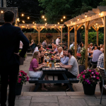 Case Study Example - Professional 16:9 hero banner. Ultra high resolution. An inviting, transformed pub garden at dusk, showcasing modern, durable outdoor furniture (recycled plastic picnic tables, stackable aluminium chairs) under warm, ambient string lights. A single, authoritative figure, possibly the pub owner, stands silhouetted confidently in the foreground, observing the bustling, happy scene. The lighting emphasizes the #737373 accent color as a subtle glow emanating from the furniture and string lights, with hints of #E2087E in the floral arrangements and #000000 in the deep shadows, creating a sophisticated, welcoming atmosphere. Style: Editorial photography with a focus on hospitality design and successful transformation.