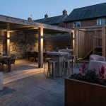 Choosing Right Pub Seating - Professional 16:9 hero banner. Ultra high resolution. An architectural detail of a stylish, modern UK pub garden, showcasing a strategically zoned outdoor area with diverse, high-quality commercial furniture. The scene features a clear distinction between a dining zone with elegant tables, a casual drinking area with high-top seating, and a family-friendly section near a secure, BS EN 1176 compliant play area. Warm, inviting accent lighting in #737373 subtly highlights the distinct zones and the durable materials, with touches of #E2087E in planters or subtle decor. The overall atmosphere is sophisticated yet welcoming, reflecting a trusted, authoritative brand. Style: Editorial architectural photography. Avoid: Generic pub scenes, people, overly bright colours, text overlays.
