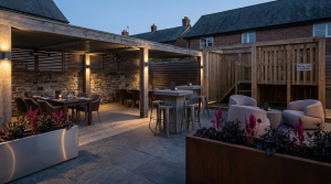 Choosing Right Pub Seating - Professional 16:9 hero banner. Ultra high resolution. An architectural detail of a stylish, modern UK pub garden, showcasing a strategically zoned outdoor area with diverse, high-quality commercial furniture. The scene features a clear distinction between a dining zone with elegant tables, a casual drinking area with high-top seating, and a family-friendly section near a secure, BS EN 1176 compliant play area. Warm, inviting accent lighting in #737373 subtly highlights the distinct zones and the durable materials, with touches of #E2087E in planters or subtle decor. The overall atmosphere is sophisticated yet welcoming, reflecting a trusted, authoritative brand. Style: Editorial architectural photography. Avoid: Generic pub scenes, people, overly bright colours, text overlays.