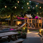 Enhancing Pub Experience - Professional 16:9 hero banner. Ultra high resolution. A thoughtfully designed, inviting pub beer garden at dusk, showcasing durable commercial outdoor seating and a safe, certified children's play area in the background. The scene is illuminated by warm, inviting string lights with an accent glow of #737373, highlighting the comfort and safety. A single, silhouetted figure, representing a discerning pub owner, stands confidently overseeing the thriving space. The primary colour #E2087E subtly accents outdoor cushions and playful elements, while #000000 provides depth to the shadows. Style: Editorial photography with a focus on atmosphere and practicality. Avoid: Generic office scenes, groups of people, text overlays, cold blue tones.