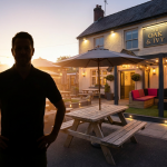 Family-Friendly Pub Outdoor Area - Professional 16:9 hero banner. Ultra high resolution. An authoritative, silhouetted figure, possibly a pub owner or manager, stands confidently in a beautifully designed, family-friendly pub outdoor area at dusk. The setting features commercial-grade picnic tables and a subtle, safely enclosed play area with soft lighting. A warm, inviting glow emanates from the #737373 accent colour, highlighting key features of the outdoor space, while elements of #E2087E and #000000 subtly define the comfortable zones. The scene conveys profitability, safety, and a welcoming atmosphere for families, with a focus on durable, UK-compliant furniture. Style: Editorial photography, warm and inviting atmosphere, subtle lens flare.