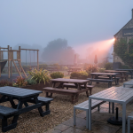 Pub Outdoor Seating - Professional 16:9 hero banner. Ultra high resolution. An evocative, slightly misty, early morning scene of a well-designed, yet empty, UK pub garden. The focus is on a strategic layout featuring robust, weatherproof pub outdoor seating (recycled plastic picnic benches and powder-coated aluminium tables) alongside a safe, EN 1176 certified children's play area with soft fall surfacing. The #737373 accent colour provides a subtle, warm glow on the polished surfaces of the furniture, hinting at potential. A single beam of light, tinged with #E2087E, cuts through the mist, illuminating the organised space. The atmosphere is quiet, authoritative, and ready for business. Style: Editorial photography with a soft, diffused light.