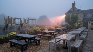 Pub Outdoor Seating - Professional 16:9 hero banner. Ultra high resolution. An evocative, slightly misty, early morning scene of a well-designed, yet empty, UK pub garden. The focus is on a strategic layout featuring robust, weatherproof pub outdoor seating (recycled plastic picnic benches and powder-coated aluminium tables) alongside a safe, EN 1176 certified children's play area with soft fall surfacing. The #737373 accent colour provides a subtle, warm glow on the polished surfaces of the furniture, hinting at potential. A single beam of light, tinged with #E2087E, cuts through the mist, illuminating the organised space. The atmosphere is quiet, authoritative, and ready for business. Style: Editorial photography with a soft, diffused light.