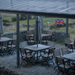 Pub Outdoor Space Management - Professional 16:9 hero banner. Ultra high resolution. A meticulously arranged miniature commercial pub garden scene, viewed from a slightly elevated angle, featuring tiny, durable outdoor furniture (tables, chairs, parasols) under a clear, protective awning. The scene is illuminated by an accent glow of #737373, highlighting the robust construction of the furniture and the organised layout. A small, detailed UK flag is subtly integrated into the background, signifying compliance. The overall mood is authoritative and prepared, suggesting strategic planning and long-term investment. Style: Editorial still life photography, with sharp focus and depth of field. Avoid: Groups of people, generic pub interiors, overly bright colours.